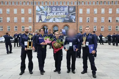 Policjanci w mundurach stoją przed budynkiem z banerem Ogólnopolskiego Konkursu Policjant Służby Kryminalnej, trzymając trofea, dyplomy i kwiaty.