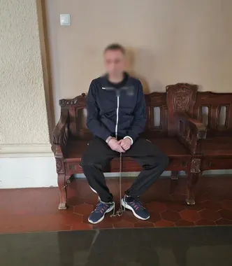 Mężczyzna w ciemnym dresie, zakuty w kajdanki i obrożę na nogach, siedzi na drewnianej ławce w pomieszczeniu o ceglastej posadzce.