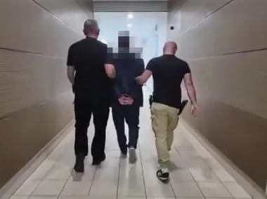 Trzech mężczyzn idzie korytarzem, przy czym dwaj z nich prowadzą trzeciego, który ma skrępowane ręce za plecami.