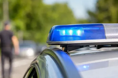 Zbliżenie na niebieski sygnalizator świetlny na dachu policyjnego radiowozu, w tle rozmyta postać funkcjonariusza i pojazd na drodze.