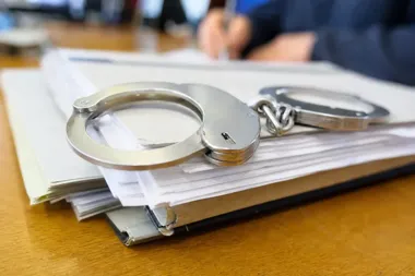 Metalowe kajdanki leżą na stosie dokumentów w teczce, na drewnianym stole; w tle widoczne ręce osoby piszącej długopisem.